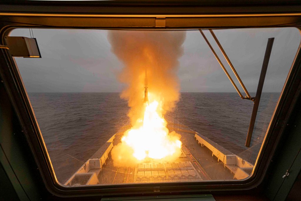 View from inside a ship's control room showing a missile launch over the ocean, with fire and smoke erupting from the missile.