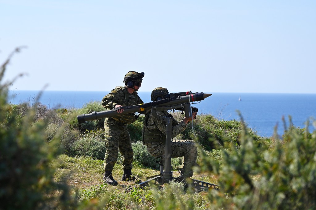 Two soldiers in military gear operating a missile launcher on a coastal landscape overlooking the sea.