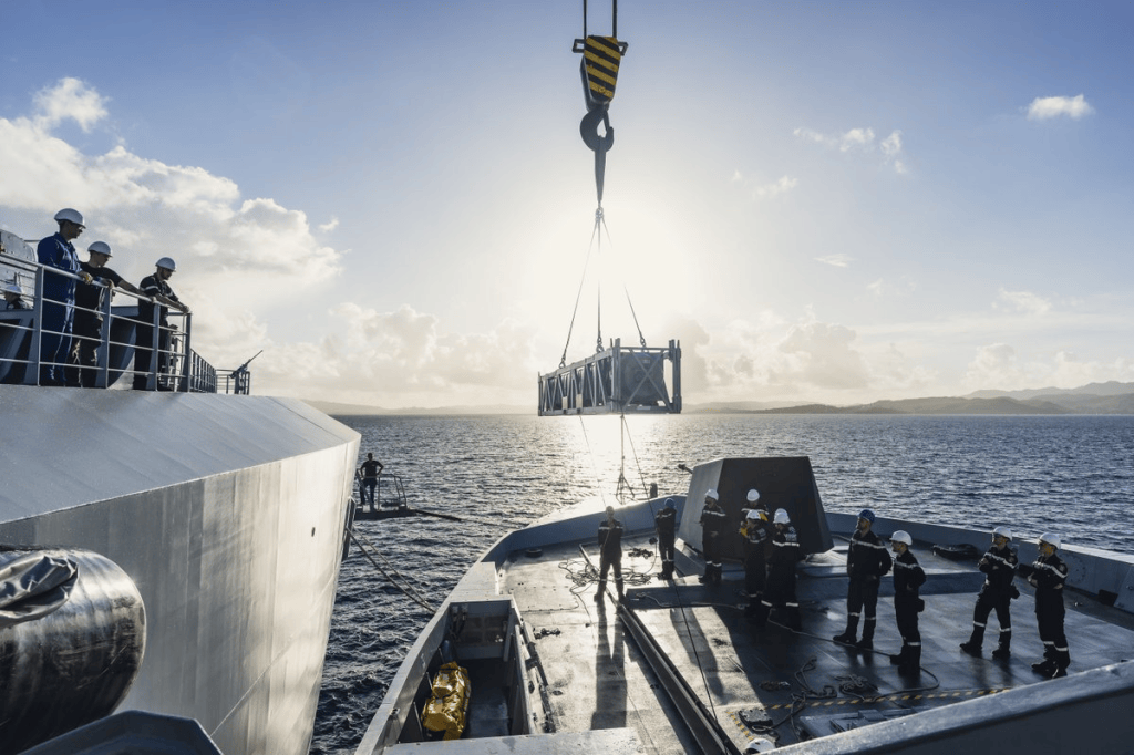 A crane is lifting a large metal structure from a ship while crew members observe from the deck, with a sunset backdrop over the ocean.