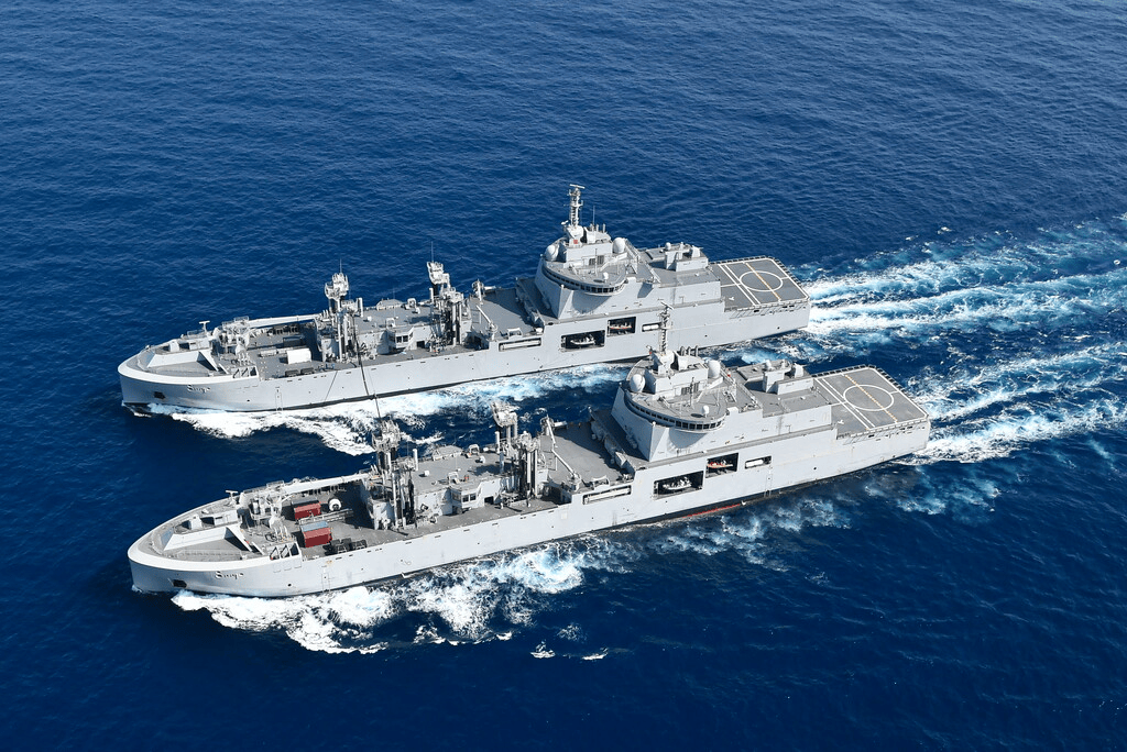 Two military vessels sailing in blue ocean waters, creating wake behind them.