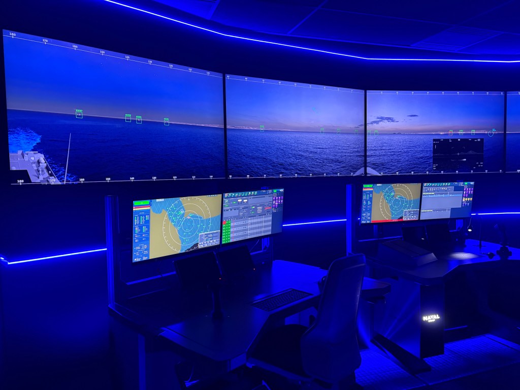 A modern maritime control room featuring multiple large screens displaying oceanic radar and navigational data, with blue ambient lighting and dual workstation setups.