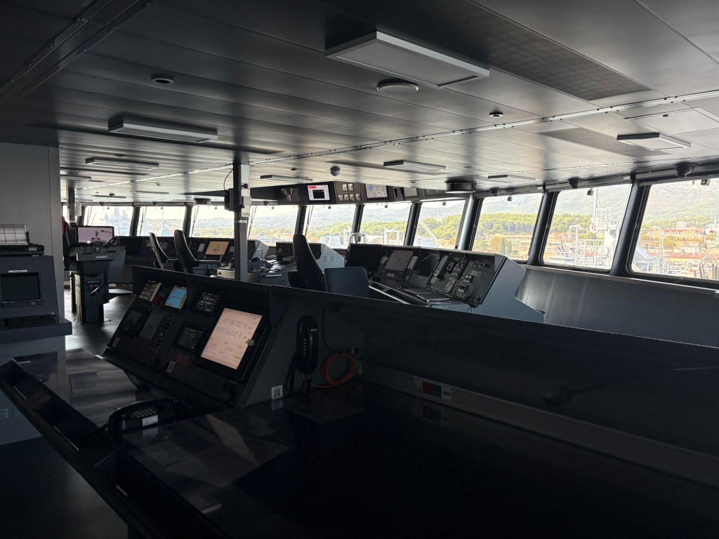 Interior view of a ship's bridge, featuring control panels, displays, and seating, with large windows showing an outdoor view.