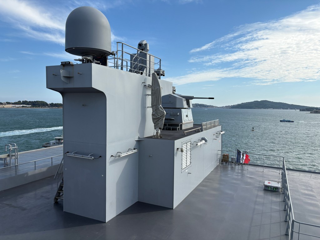A close-up view of a naval ship's upper deck featuring a radar system, a mounted gun, and a crew member, with a scenic waterfront backdrop.