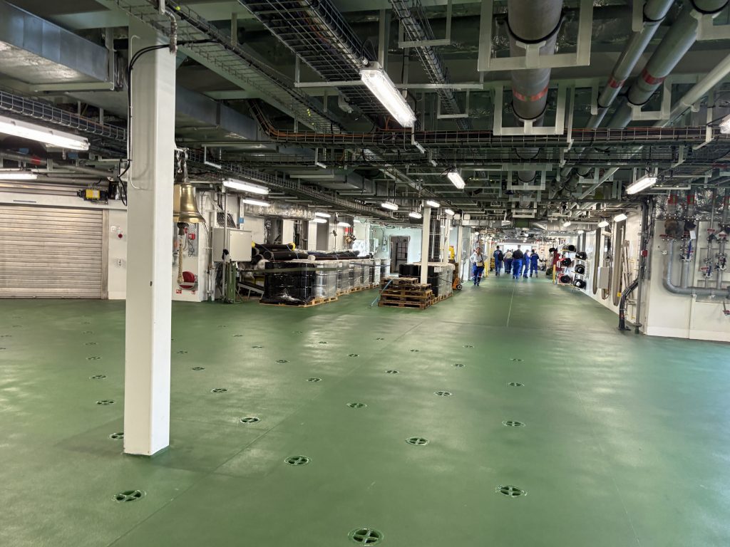 Interior view of a large industrial space with green flooring, featuring pipes, equipment, and a group of people in blue uniforms in the background.